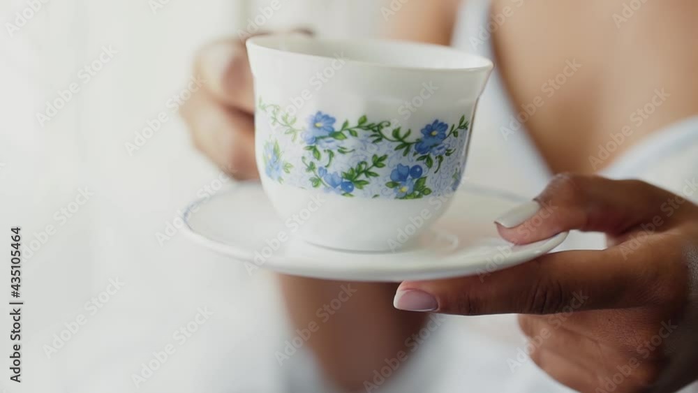 Dark skin woman holds white saucer with cup. On the mug is floral drawing with forget-me-nots. The african woman raises her cup in morning. Dark skin woman holds white saucer with cup. On the mug is floral drawing with forget-me-nots. The african woman raises her cup in morning.