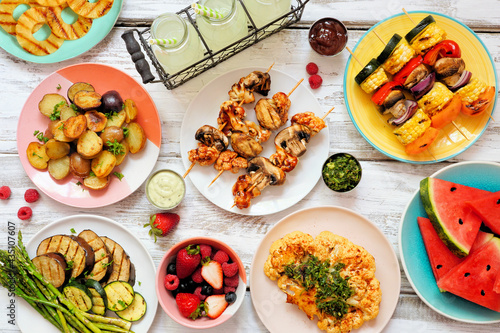 Vegan summer bbq or picnic table scene. Overhead view on a white wood background. Fruit, grilled vegetables, skewers, cauliflower steak and lemonade. Meat substitute concept.