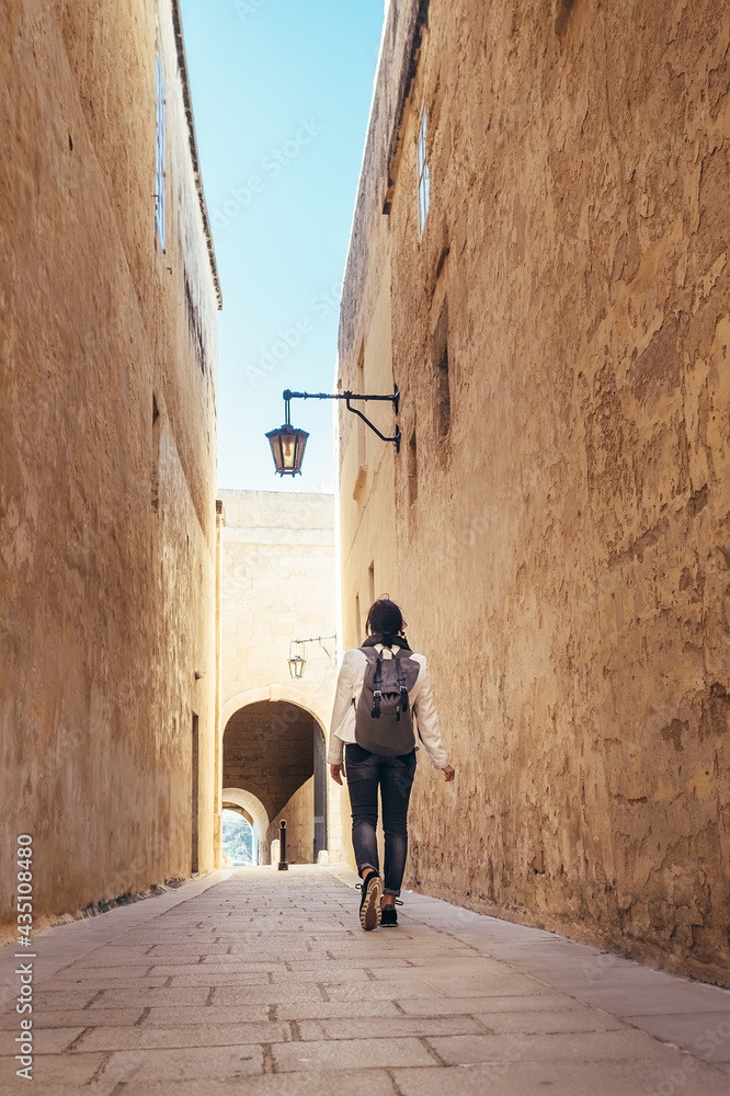 Female traveling alone on the Malta island. Tourist walking by the narrow ancient medieval Mdina streets with a city backpack and enjoying an historical place.