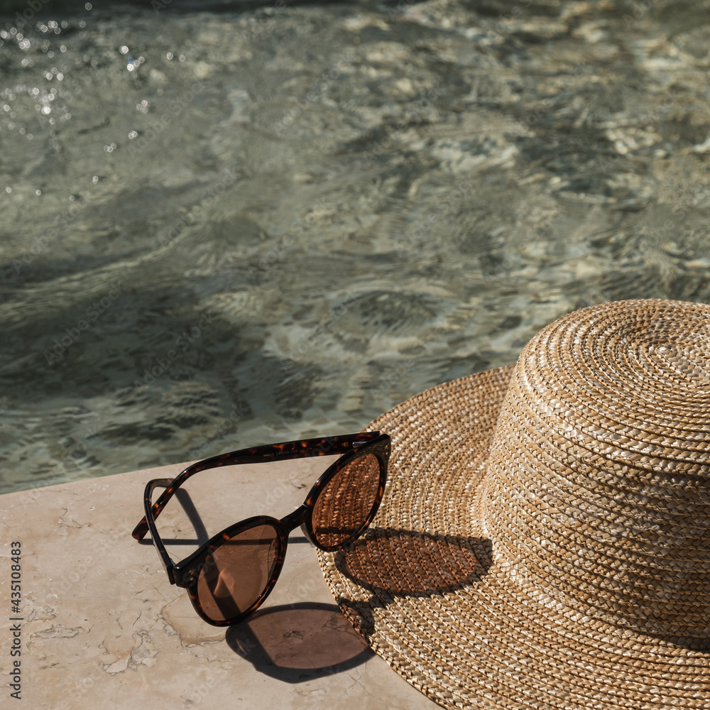 Sunglasses and straw hat on marble swimming pool side with clear blue water  with waves sunlight shadow reflections. Minimal fashion aesthetic summer  vacation creative background Stock Photo