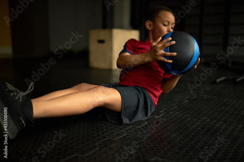 Active self determined little African boy wearing sports clothing and sneakers doing sit ups abdominal crunches for core muscles using heavy medicine ball having tired facial expression photo