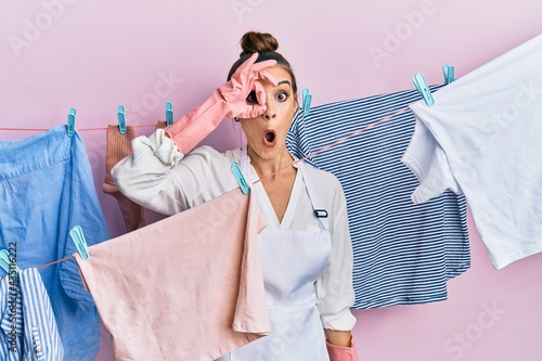 Beautiful brunette young woman washing clothes at clothesline doing ok gesture shocked with surprised face, eye looking through fingers. unbelieving expression.