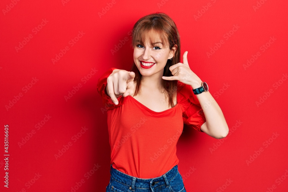 Redhead young woman wearing casual red t shirt smiling doing talking on the telephone gesture and pointing to you. call me. Redhead young woman wearing casual red t shirt smiling doing talking on the telephone gesture and pointing to you. call me.