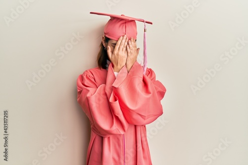 Young caucasian woman wearing graduation cap and ceremony robe with sad expression covering face with hands while crying. depression concept.