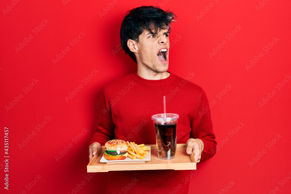 Handsome hipster young man eating a tasty classic burger with fries and soda angry and mad screaming frustrated and furious, shouting with anger. rage and aggressive concept. Handsome hipster young man eating a tasty classic burger with fries and soda angry and mad screaming frustrated and furious, shouting with anger. rage and aggressive concept.
