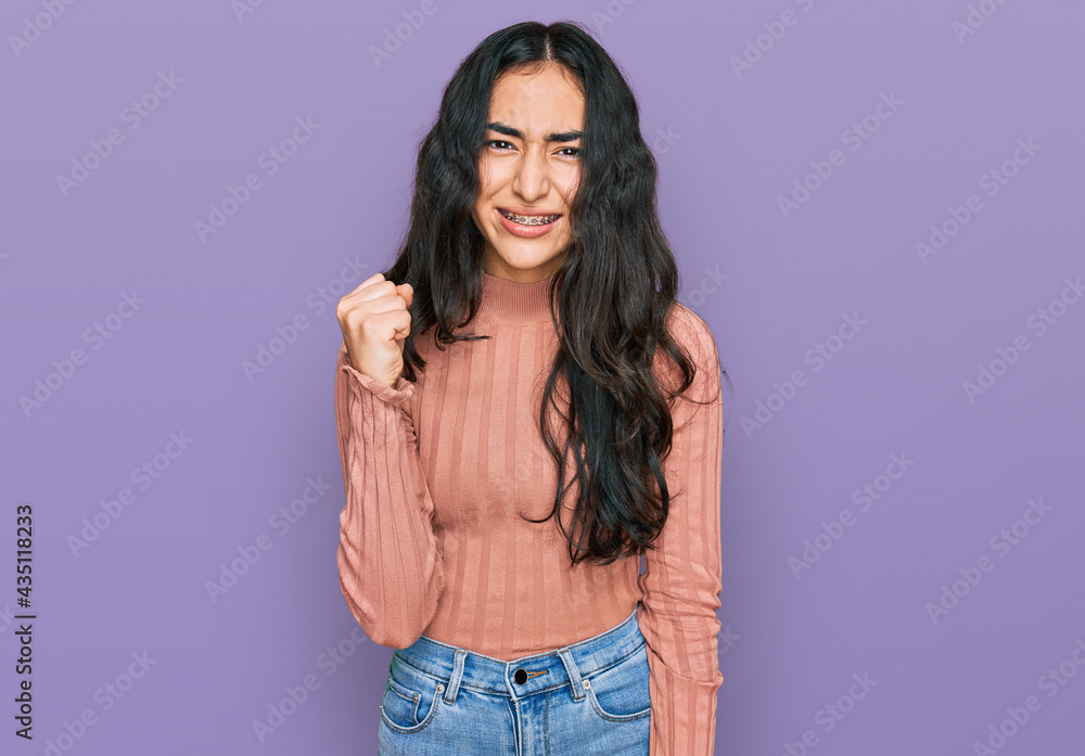 Hispanic teenager girl with dental braces wearing casual clothes angry and mad raising fist frustrated and furious while shouting with anger. rage and aggressive concept. Hispanic teenager girl with dental braces wearing casual clothes angry and mad raising fist frustrated and furious while shouting with anger. rage and aggressive concept.