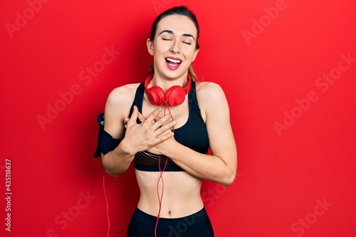 Young hispanic girl wearing gym clothes and using headphones smiling with hands on chest with closed eyes and grateful gesture on face. health concept.