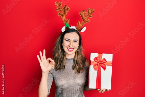 Young hispanic girl wearing deer christmas hat holding gift doing ok sign with fingers, smiling friendly gesturing excellent symbol