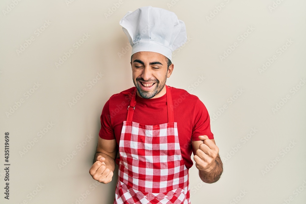 Young hispanic man wearing baker uniform very happy and excited doing winner gesture with arms raised, smiling and screaming for success. celebration concept. Young hispanic man wearing baker uniform very happy and excited doing winner gesture with arms raised, smiling and screaming for success. celebration concept.