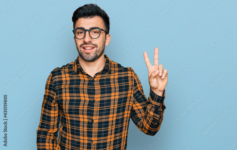 Hispanic man with beard wearing casual clothes and glasses smiling with happy face winking at the camera doing victory sign. number two. Hispanic man with beard wearing casual clothes and glasses smiling with happy face winking at the camera doing victory sign. number two.