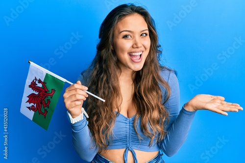 Young hispanic girl holding wales flag celebrating achievement with happy smile and winner expression with raised hand