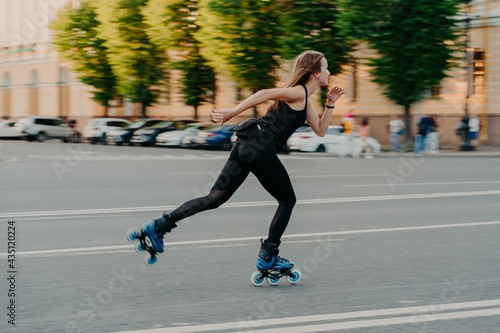Canvas-taulu Professional female roller demonstrates her abilities of rollerblading rides very quckly on road along city enjoys sunny day dressed in black active wear