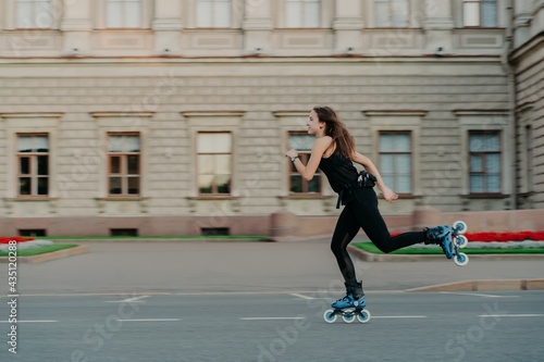 Outdoor physical activities. Healthy young woman has slim fit figure leads active lifestyle spends free time outside rides on rollerblades poses alone in open air against building background