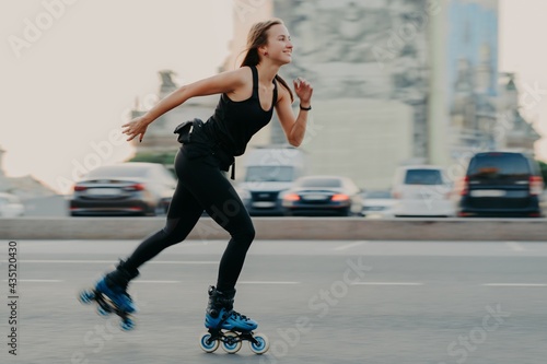 Young dark haired slim healthy woman has recration time enjoys rolleblading on speed moves fast has cheerful expression wears black t shirt and leggings. Active rest and sport training concept