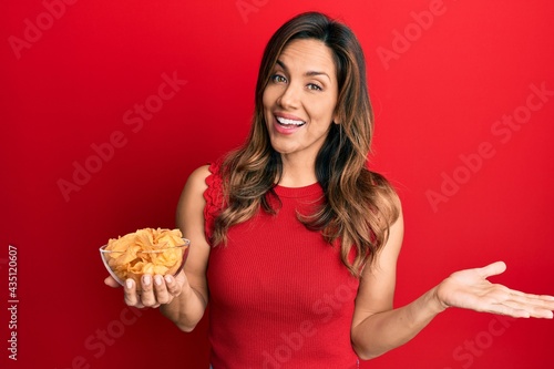 Young latin woman holding potato chip celebrating achievement with happy smile and winner expression with raised hand