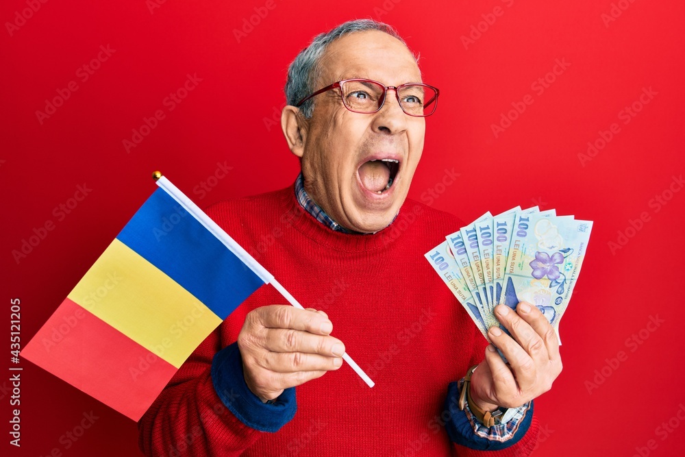 Handsome senior man with grey hair holding romania flag and leu banknotes angry and mad screaming frustrated and furious, shouting with anger. rage and aggressive concept. Handsome senior man with grey hair holding romania flag and leu banknotes angry and mad screaming frustrated and furious, shouting with anger. rage and aggressive concept.