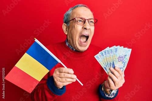 Handsome senior man with grey hair holding romania flag and leu banknotes angry and mad screaming frustrated and furious, shouting with anger. rage and aggressive concept.