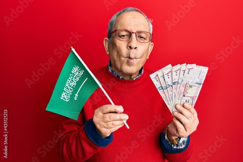 Handsome senior man with grey hair holding saudi arabia flag and riyal banknotes making fish face with mouth and squinting eyes, crazy and comical.