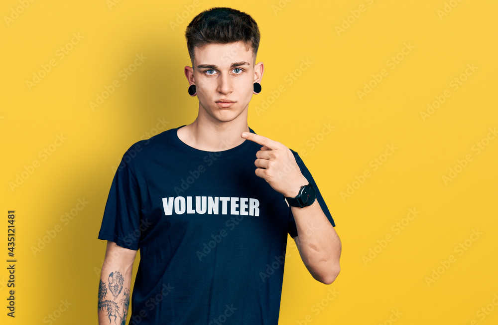 Young caucasian boy with ears dilation wearing volunteer t shirt pointing with hand finger to the side showing advertisement, serious and calm face Young caucasian boy with ears dilation wearing volunteer t shirt pointing with hand finger to the side showing advertisement, serious and calm face