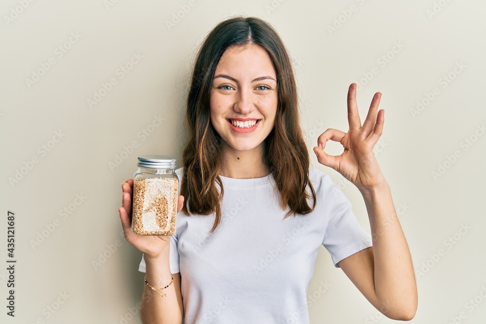 Young brunette woman holding jar with rice doing ok sign with fingers, smiling friendly gesturing excellent symbol Young brunette woman holding jar with rice doing ok sign with fingers, smiling friendly gesturing excellent symbol