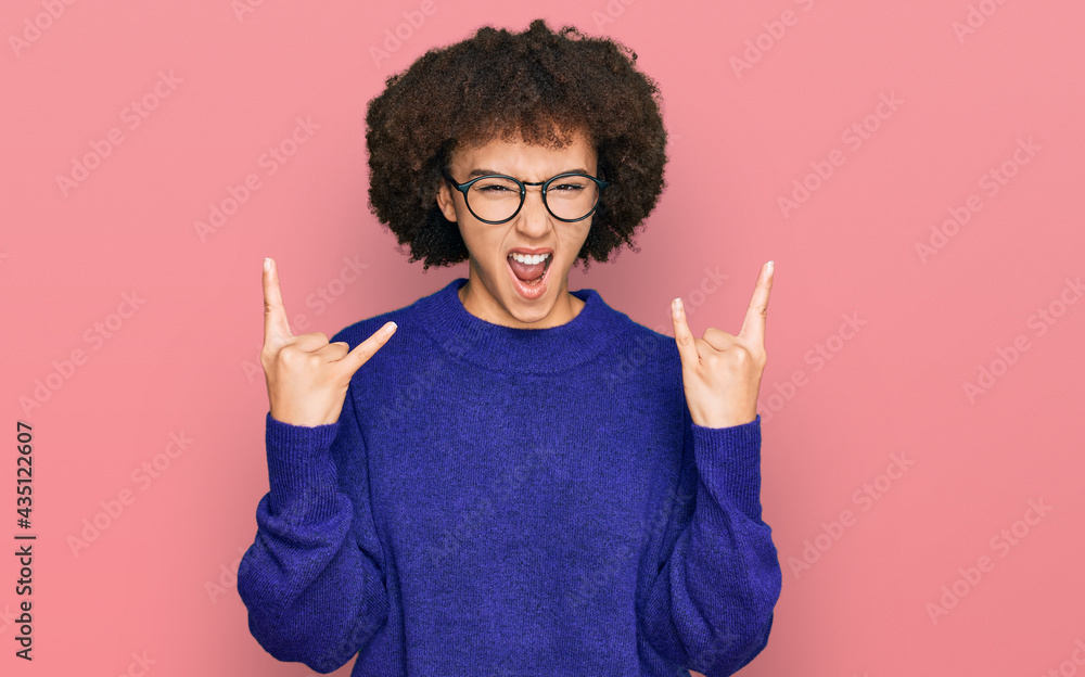 Young hispanic girl wearing casual winter sweater and glasses shouting with crazy expression doing rock symbol with hands up. music star. heavy concept. Young hispanic girl wearing casual winter sweater and glasses shouting with crazy expression doing rock symbol with hands up. music star. heavy concept.