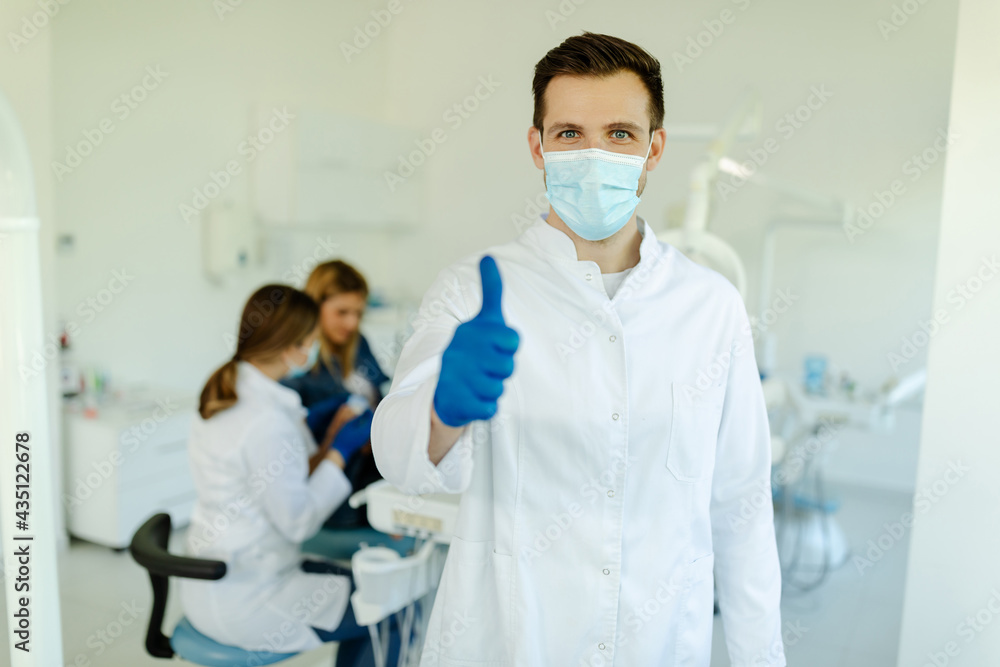 Smiling male dentist wearing mask and gloves showing thumbs up over medical office background. Smiling male dentist wearing mask and gloves showing thumbs up over medical office background.