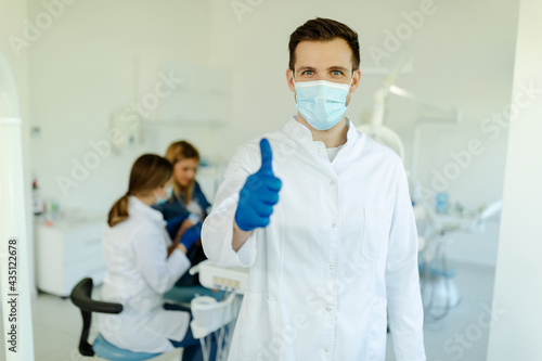 Smiling male dentist wearing mask and gloves showing thumbs up over medical office background.
