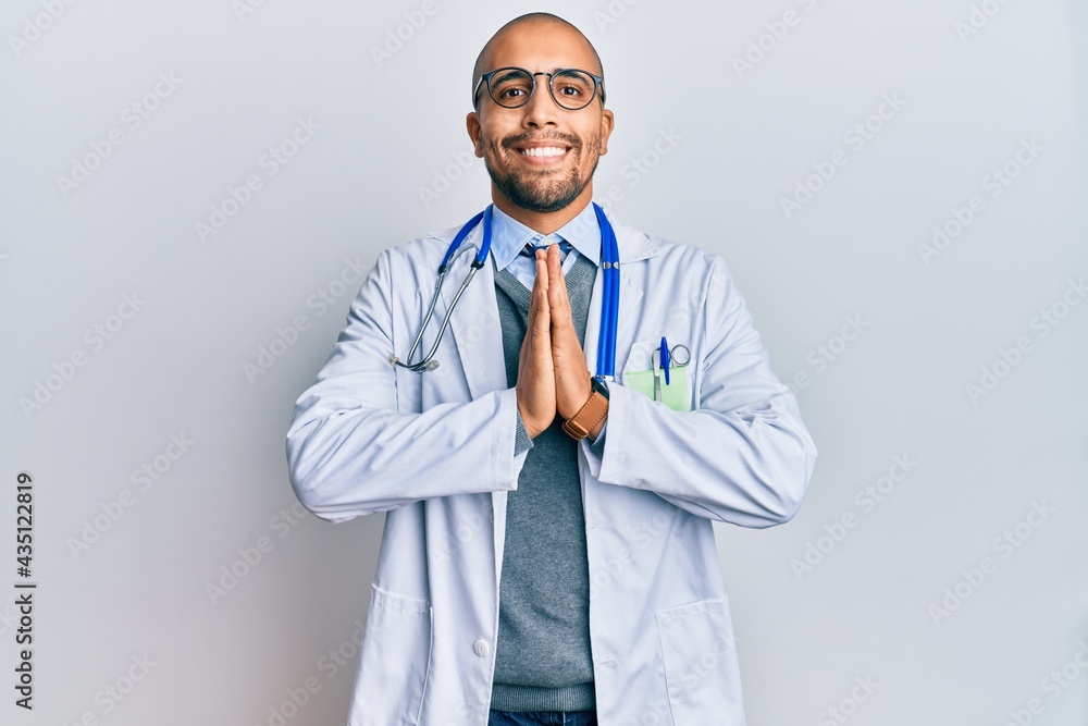 Hispanic adult man wearing doctor uniform and stethoscope praying with hands together asking for forgiveness smiling confident. Hispanic adult man wearing doctor uniform and stethoscope praying with hands together asking for forgiveness smiling confident.