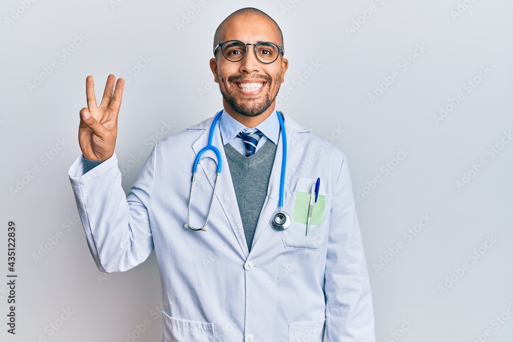Hispanic adult man wearing doctor uniform and stethoscope showing and pointing up with fingers number three while smiling confident and happy.