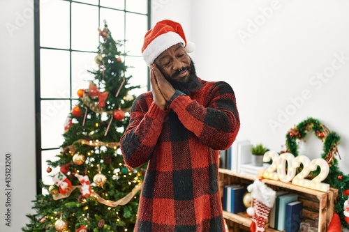 African american man wearing santa claus hat standing by christmas tree sleeping tired dreaming and posing with hands together while smiling with closed eyes.