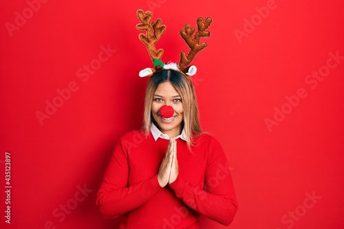 Beautiful hispanic woman wearing deer christmas hat and red nose praying with hands together asking for forgiveness smiling confident.