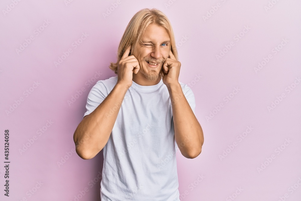 Caucasian young man with long hair wearing casual white t shirt covering ears with fingers with annoyed expression for the noise of loud music. deaf concept. Caucasian young man with long hair wearing casual white t shirt covering ears with fingers with annoyed expression for the noise of loud music. deaf concept.