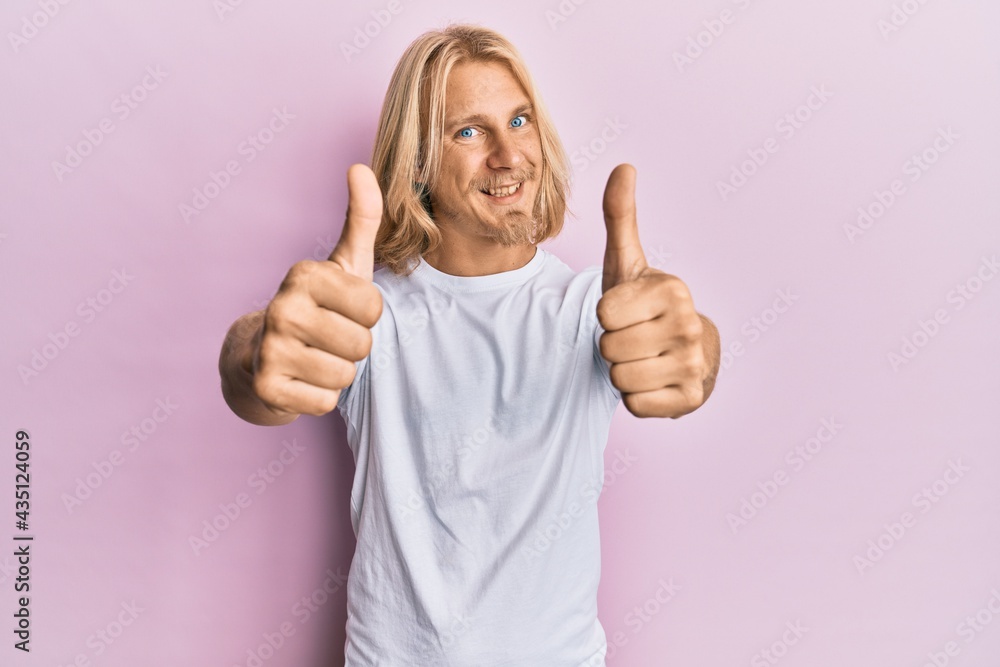 Caucasian young man with long hair wearing casual white t shirt approving doing positive gesture with hand, thumbs up smiling and happy for success. winner gesture. Caucasian young man with long hair wearing casual white t shirt approving doing positive gesture with hand, thumbs up smiling and happy for success. winner gesture.
