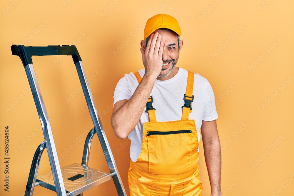 Handsome middle age man with grey hair holding ladder covering one eye with hand, confident smile on face and surprise emotion. Handsome middle age man with grey hair holding ladder covering one eye with hand, confident smile on face and surprise emotion.