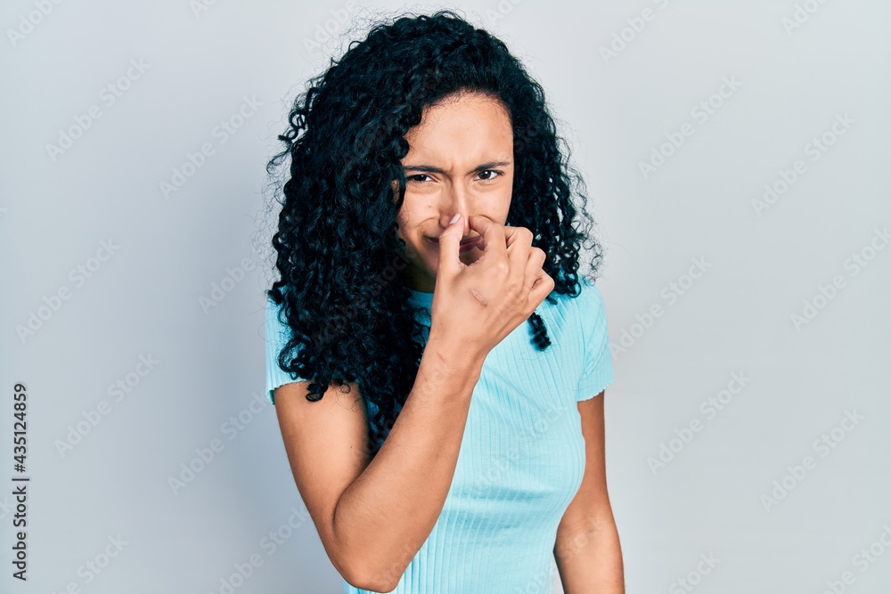 Young hispanic woman with curly hair wearing casual blue t shirt smelling something stinky and disgusting, intolerable smell, holding breath with fingers on nose. bad smell Young hispanic woman with curly hair wearing casual blue t shirt smelling something stinky and disgusting, intolerable smell, holding breath with fingers on nose. bad smell