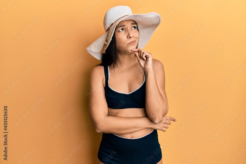 Young latin woman wearing bikini and summer hat serious face thinking about question with hand on chin, thoughtful about confusing idea Young latin woman wearing bikini and summer hat serious face thinking about question with hand on chin, thoughtful about confusing idea