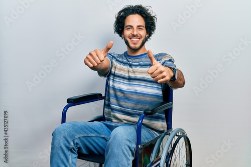 Handsome hispanic man sitting on wheelchair approving doing positive gesture with hand, thumbs up smiling and happy for success. winner gesture.