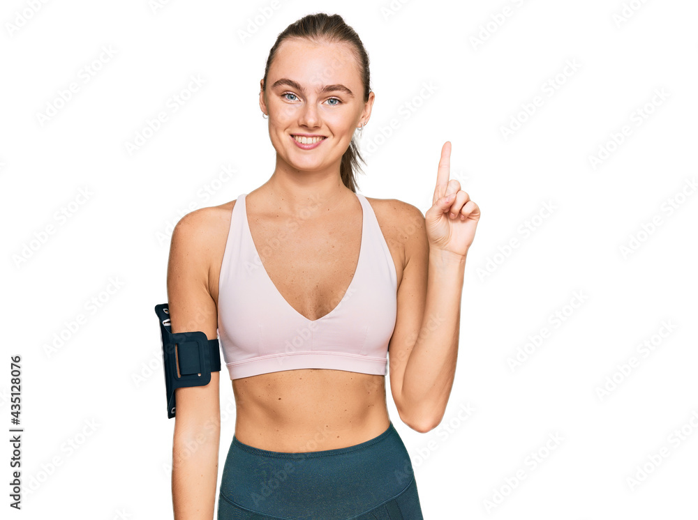 Beautiful young blonde woman wearing sportswear and arm band pointing finger up with successful idea. exited and happy. number one. Beautiful young blonde woman wearing sportswear and arm band pointing finger up with successful idea. exited and happy. number one.