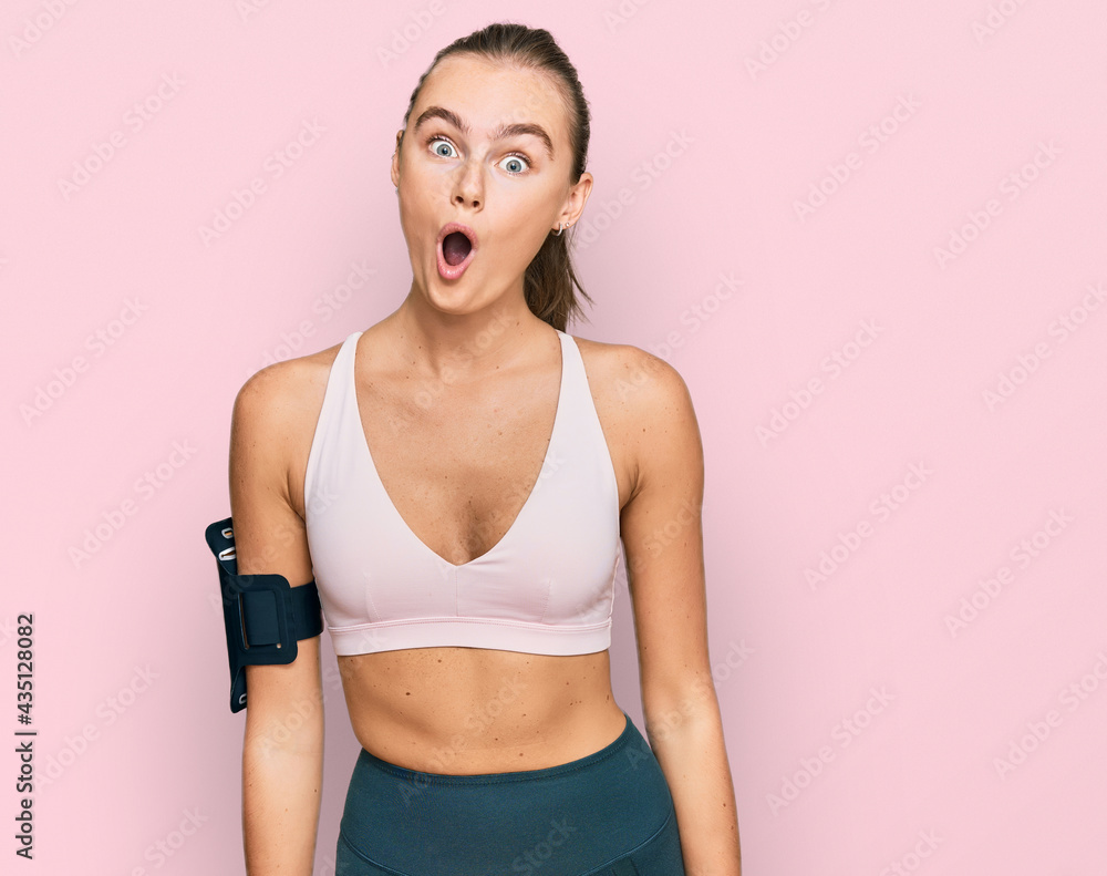 Beautiful young blonde woman wearing sportswear and arm band scared and amazed with open mouth for surprise, disbelief face Beautiful young blonde woman wearing sportswear and arm band scared and amazed with open mouth for surprise, disbelief face