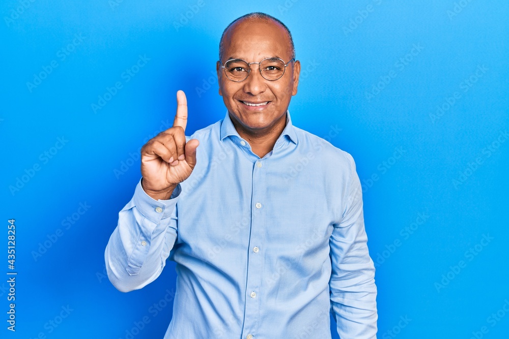 Middle age latin man wearing casual clothes and glasses showing and pointing up with finger number one while smiling confident and happy. Middle age latin man wearing casual clothes and glasses showing and pointing up with finger number one while smiling confident and happy.