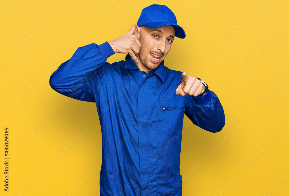 Bald man with beard wearing builder jumpsuit uniform smiling doing talking on the telephone gesture and pointing to you. call me.