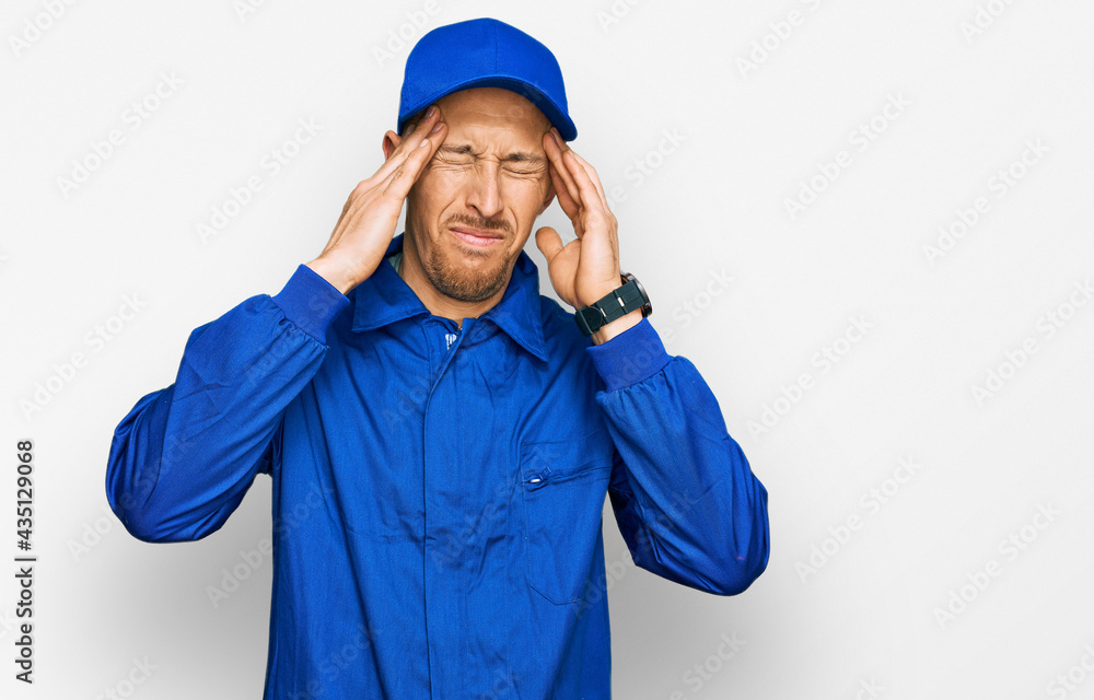 Bald man with beard wearing builder jumpsuit uniform with hand on head, headache because stress. suffering migraine. Bald man with beard wearing builder jumpsuit uniform with hand on head, headache because stress. suffering migraine.