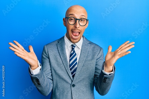 Bald man with beard wearing business jacket and glasses celebrating victory with happy smile and winner expression with raised hands