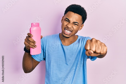 Young african american man holding shampoo bottle annoyed and frustrated shouting with anger, yelling crazy with anger and hand raised