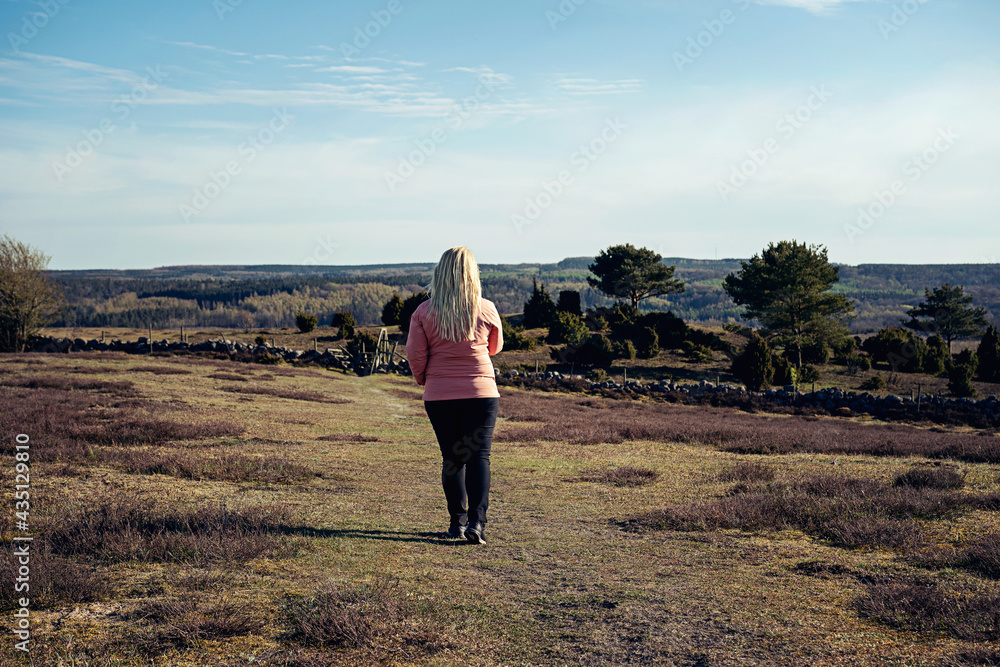 Woman with blonde long hair in pink windbreaker jacket and outdoor clothing hiking on trail through fields and beautiful landscape on a sunny day in south of Sweden. Photo taken in Brösarp, Sweden