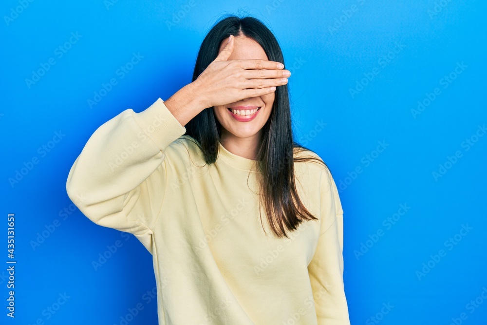 Young brunette woman wearing casual sweatshirt smiling and laughing with hand on face covering eyes for surprise. blind concept. Young brunette woman wearing casual sweatshirt smiling and laughing with hand on face covering eyes for surprise. blind concept.