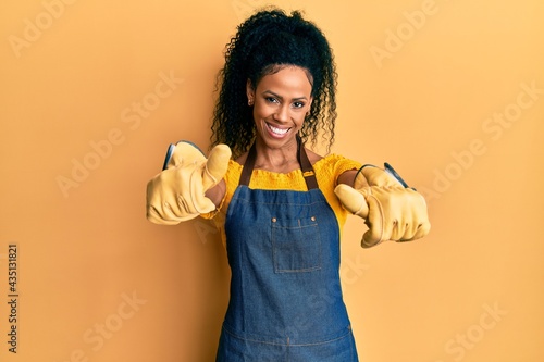 Middle age african american woman wearing professional apron approving doing positive gesture with hand, thumbs up smiling and happy for success. winner gesture.