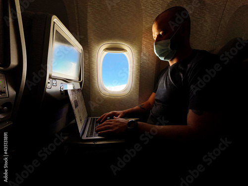Man wearing face mask working on his laptop while flying in the airplane