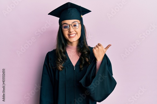 Young hispanic woman wearing graduation cap and ceremony robe smiling with happy face looking and pointing to the side with thumb up.