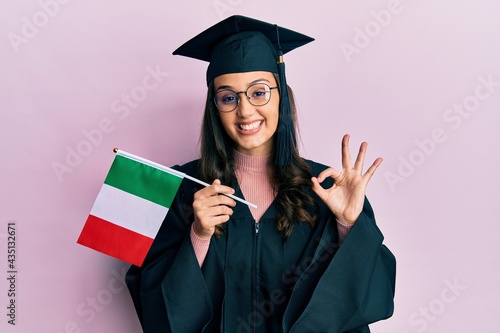 Young hispanic woman wearing graduation uniform holding italy flag doing ok sign with fingers, smiling friendly gesturing excellent symbol